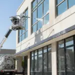 Exterior facade cleaning service worker using a hydraulic lift to power wash large glass windows of a modern commercial building in daylight.